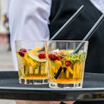 Delicious orange cocktails with cranberries and herbs on a tray, served by waiter at an outdoor event.
